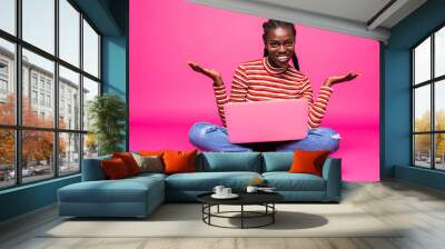 Optimistic woman with beautiful smile using silver notebook, while sitting in lotus pose on the floor and throwing hands aside isolated over pink background Wall mural